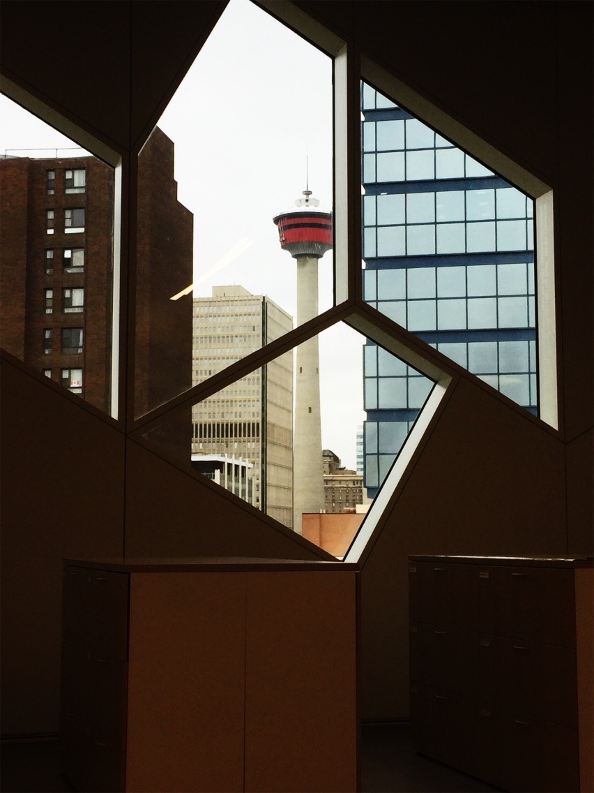 The view from the inside a dimly lit building with geometrically shaped windows that show a slim tower with a red top deck and a concrete neck that is surrounded by high rise buildings.