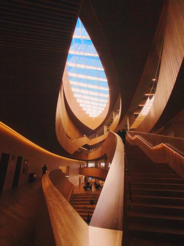 Inside a building with curved wooden architecture with wooden staircases and an ellipse shaped skylight window.